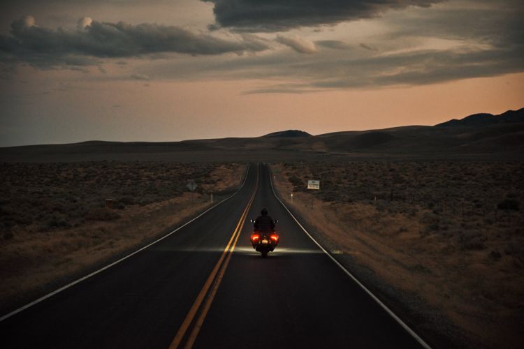 Motorcycle rider traveling down a remote highway at dusk, heading towards distant hills under a dramatic, cloudy sky.
