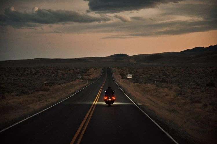 Motorcycle rider traveling down a remote highway at dusk, heading towards distant hills under a dramatic, cloudy sky.