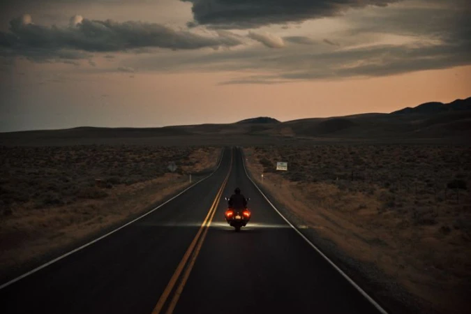 Motorcycle rider traveling down a remote highway at dusk, heading towards distant hills under a dramatic, cloudy sky.