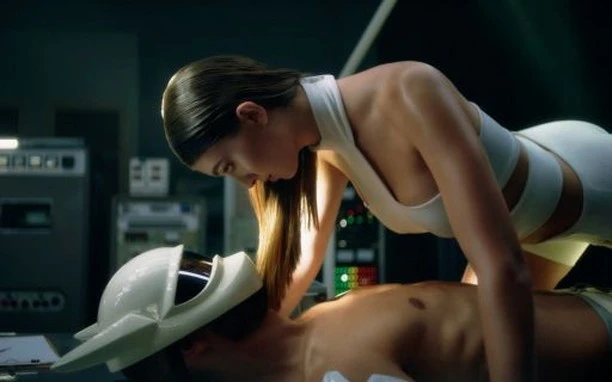 Adult woman in futuristic outfit leans over a man wearing a helmet on a table in an indoor, laboratory-like setting with equipment.