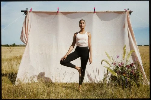 Woman in pink tank top and black leggings balancing in tree pose before a fabric backdrop outdoors in a field