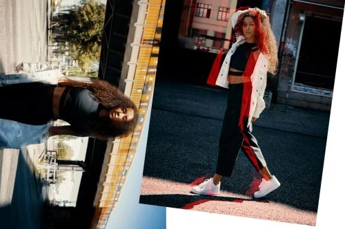 Young woman in a black crop top and pants with a white jacket with orange and red hair poses outside in an urban setting.