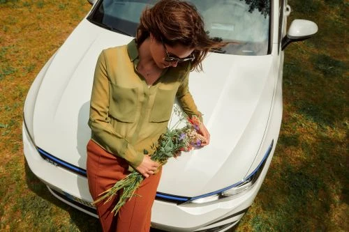 Woman in olive shirt with flowers leans on a white car on a grassy field, taken from a high angle.