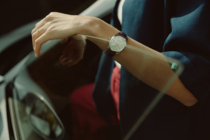 Close up of a person's arm with a wristwatch, visible through a car window. The person wears a navy blazer and a ring.