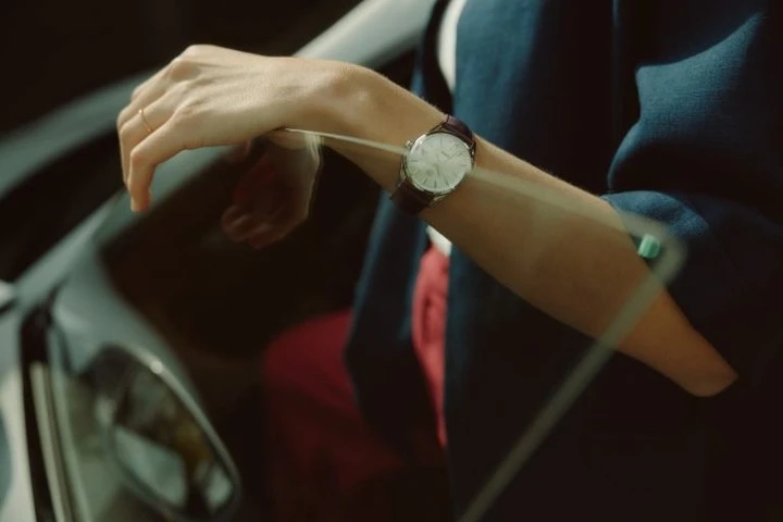Close up of a person's arm with a wristwatch, visible through a car window. The person wears a navy blazer and a ring.