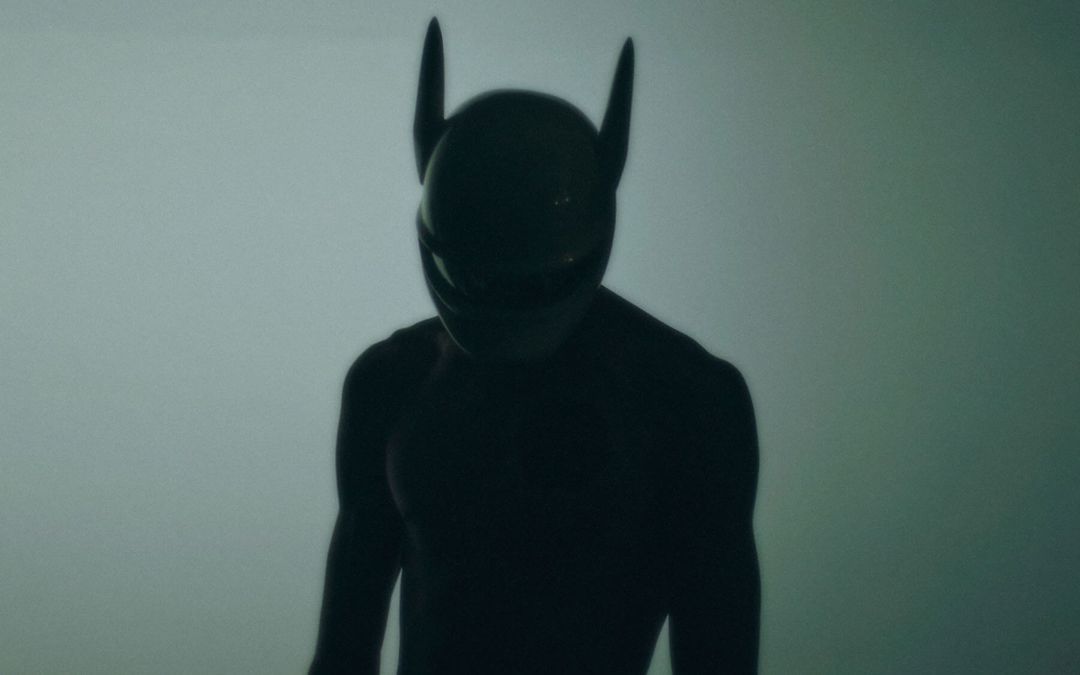 Silhouette of a figure wearing a black helmet with horn-like protrusions against a muted background, obscured by shadow.