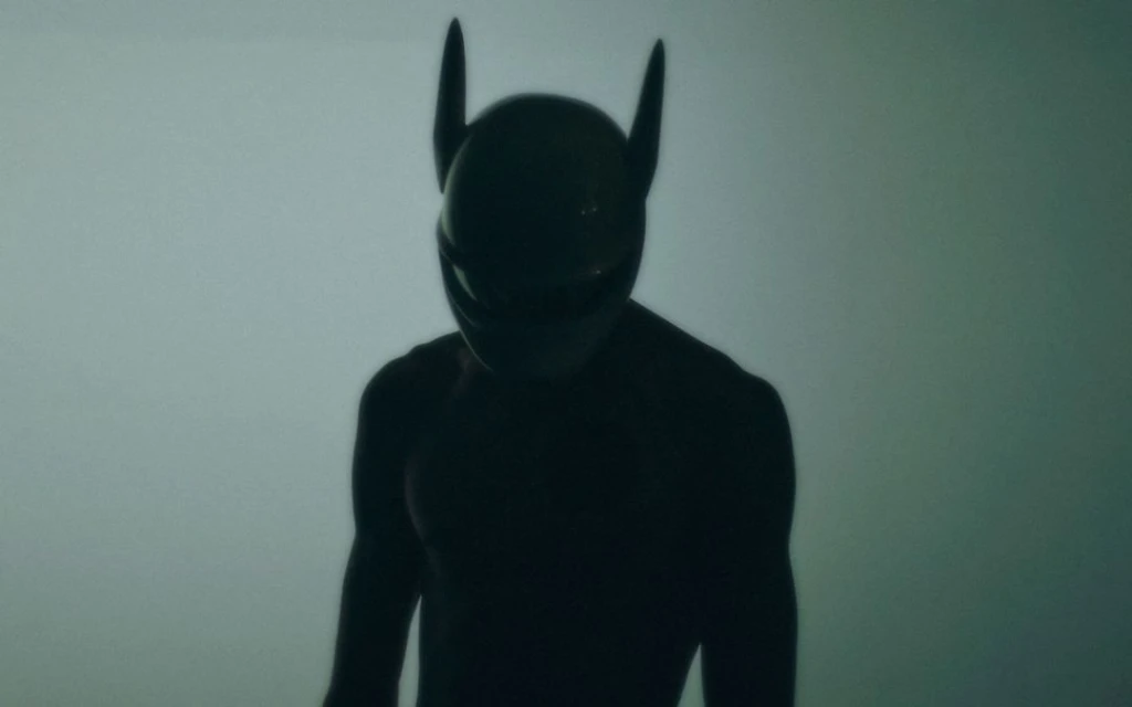 Silhouette of a figure wearing a black helmet with horn-like protrusions against a muted background, obscured by shadow.