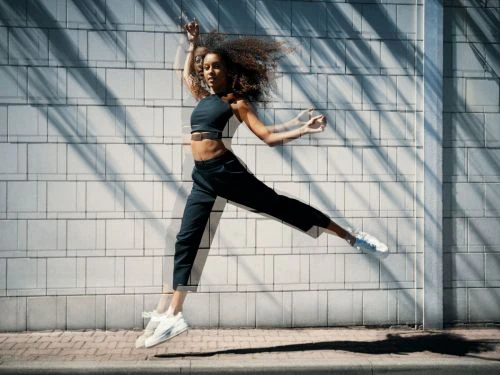 Athletic woman with curly hair in dynamic mid-air leap against a textured wall with dappled light, wearing sportswear and sneakers.