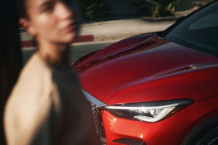Close-up of a red Infiniti car with an out-of-focus woman in the background, captured during daylight in an outdoor setting.