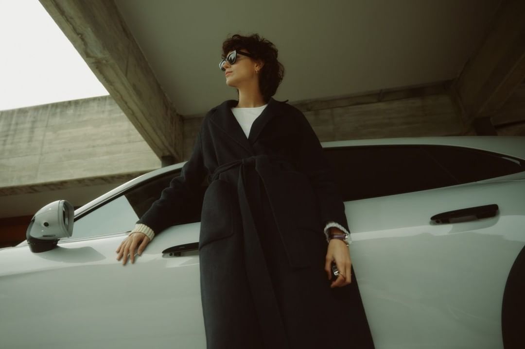 A person wearing a dark coat and sunglasses leans against a white car under a concrete structure, photographed by David Daub.