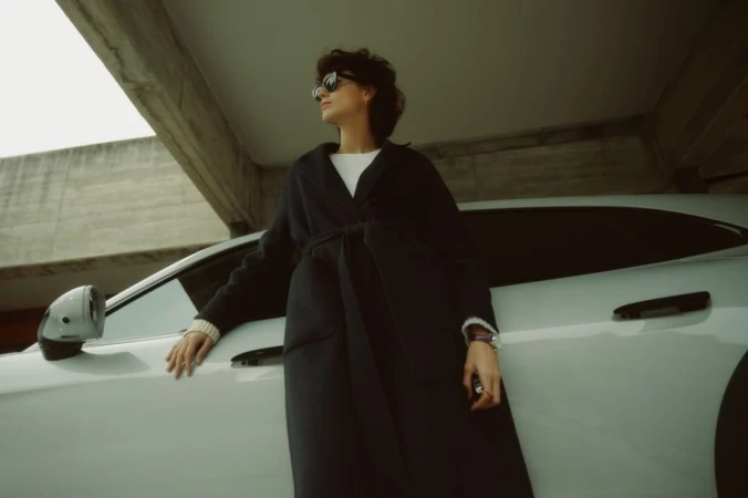 A person wearing a dark coat and sunglasses leans against a white car under a concrete structure, photographed by David Daub.