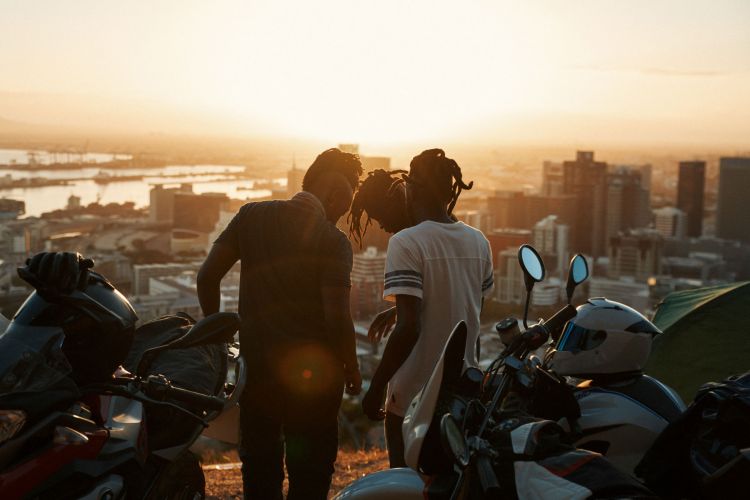 Three people silhouetted against a sunset overlooking a city, with motorcycles parked in the foreground, creating a scenic, urban landscape.