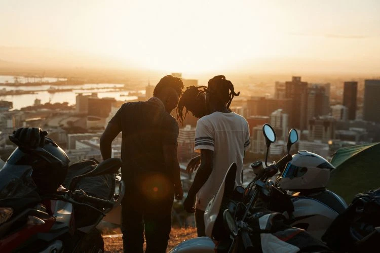 Three people silhouetted against a sunset overlooking a city, with motorcycles parked in the foreground, creating a scenic, urban landscape.