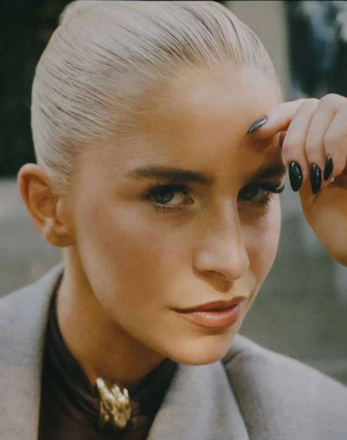 Close-up portrait of a blonde woman with green eyes and dark nails wearing a grey jacket and jewelry, looking directly at the camera.
