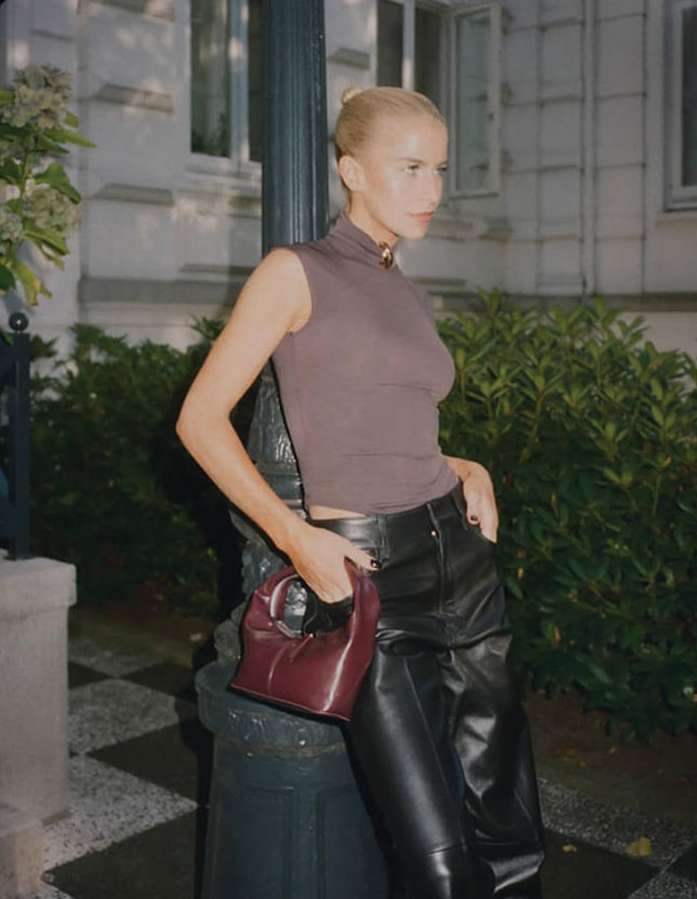 A woman in leather pants poses outdoors, leaning on a pole while holding a purse, creating a stylish fashion portrait.
