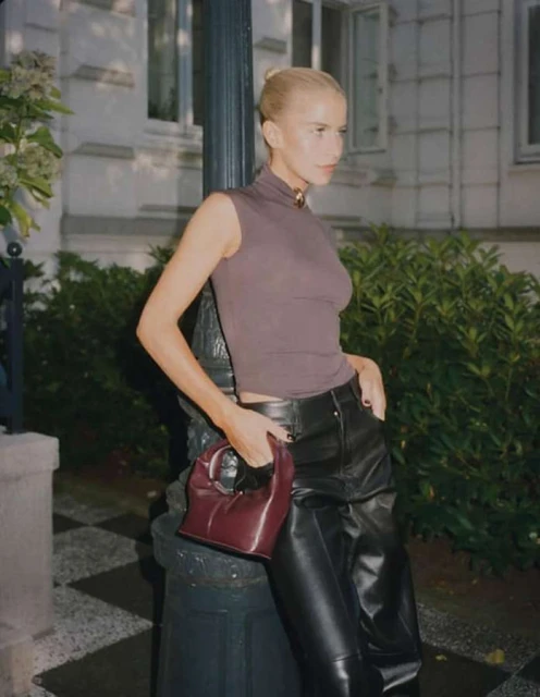 A woman in leather pants poses outdoors, leaning on a pole while holding a purse, creating a stylish fashion portrait.