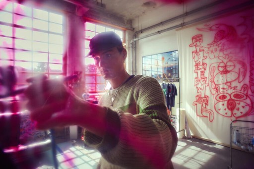 Young adult adjusts a camera in an indoor studio setting with large windows, wearing a baseball cap and casual attire.