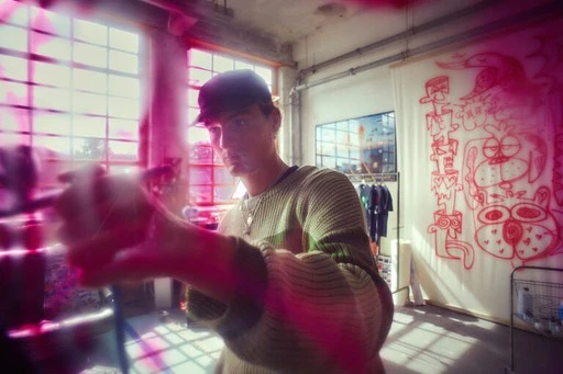 Young adult adjusts a camera in an indoor studio setting with large windows, wearing a baseball cap and casual attire.