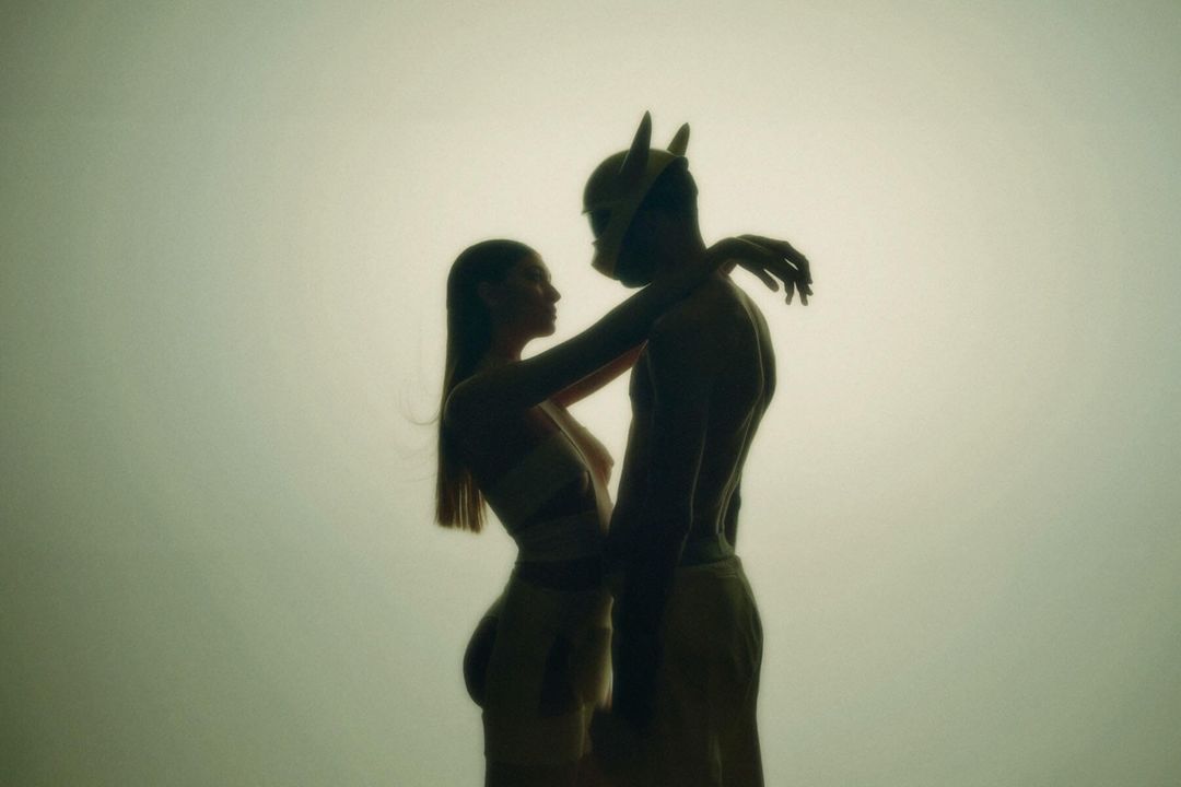 Silhouette of a shirtless male figure with horns, embracing a woman in a matching outfit against a light background.