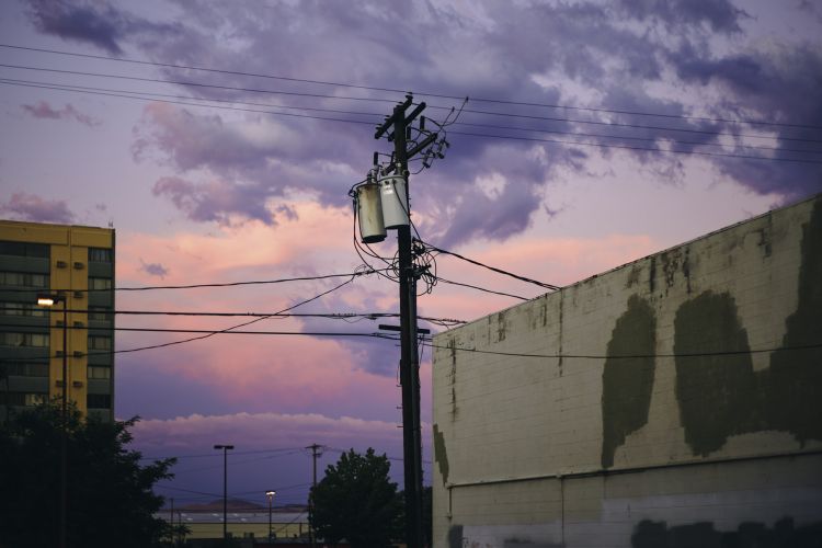 Urban landscape featuring a weathered utility pole against a sunset sky with light clouds, adjacent to a building and power lines.