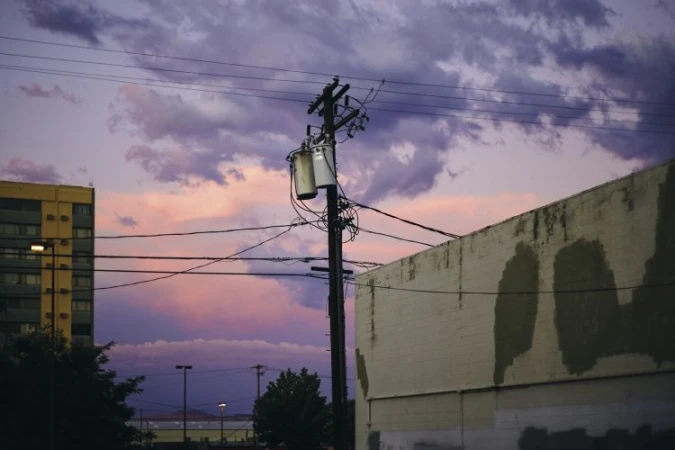 Urban landscape featuring a weathered utility pole against a sunset sky with light clouds, adjacent to a building and power lines.