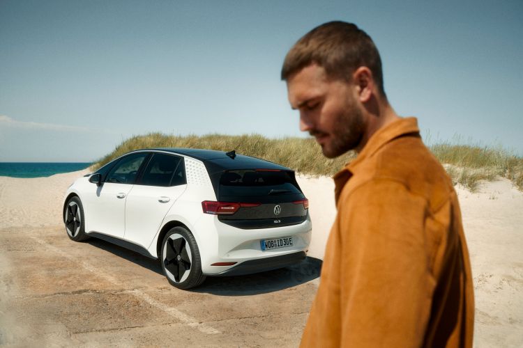 Man in amber jacket stands near a white Volkswagen ID.3 electric car on a beach with blue ocean and sky in the background on a sunny day.