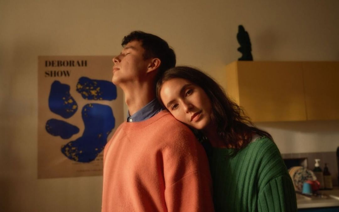 Young couple embraces indoors, woman resting head on man's shoulder, soft lighting, casual clothing, poster art in background.