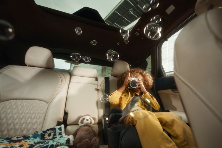 Child taking photo inside a car with bubbles, teddy bear, and a blanket. Cream leather interior, sunroof, and natural lighting.