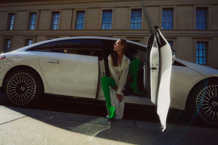 An adult female model sitting in a white Mercedes car in an urban setting, with the car door open. She is wearing green boots.