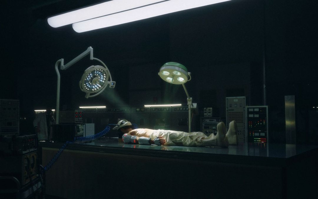 Man lies on an operating table in a dimly lit room, lit by surgical lights. Medical equipment and dark walls in the background.