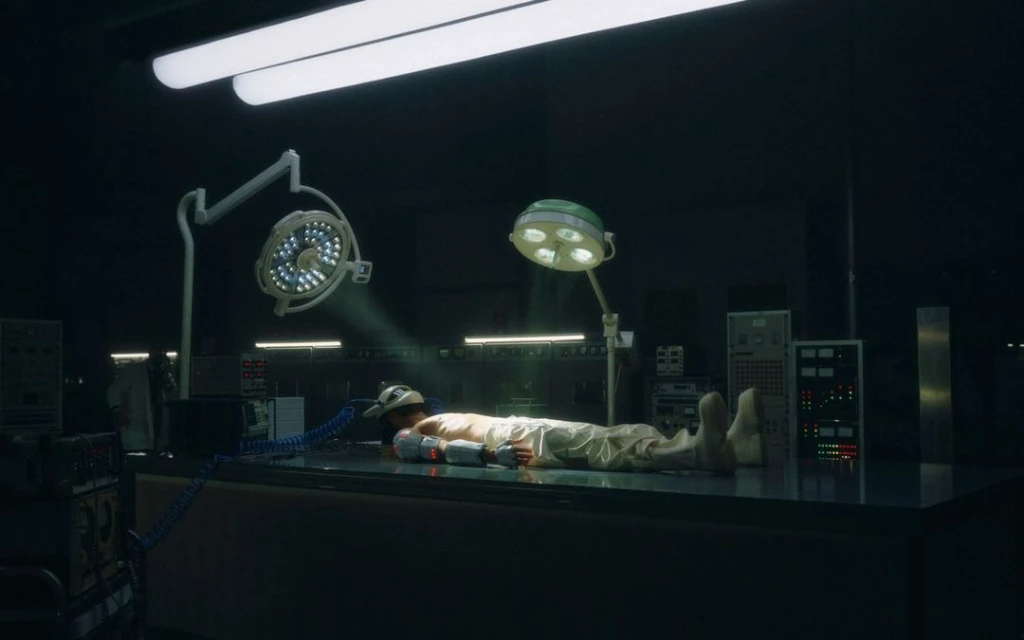 Man lies on an operating table in a dimly lit room, lit by surgical lights. Medical equipment and dark walls in the background.