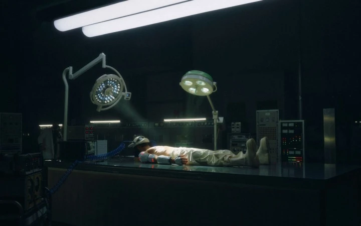 Man lies on an operating table in a dimly lit room, lit by surgical lights. Medical equipment and dark walls in the background.