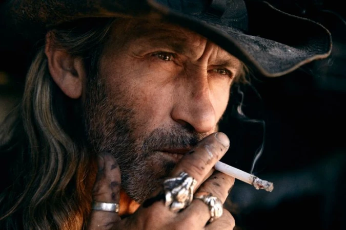 Close-up portrait of an older man with long hair, beard, cowboy hat, smoking a cigarette and wearing rings, by David Daub.