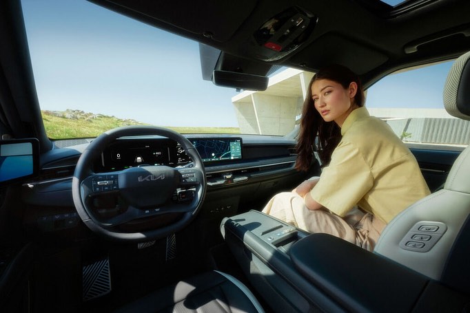Interior view of a Kia car with a woman in the passenger seat, showcasing the dashboard, steering wheel, and modern design elements.