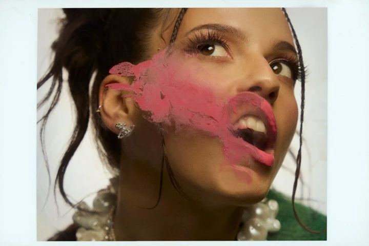 Portrait of a young woman with pink paint on her face, jewelry, and an open mouth, set against a neutral studio backdrop for a beauty or fashion campaign.