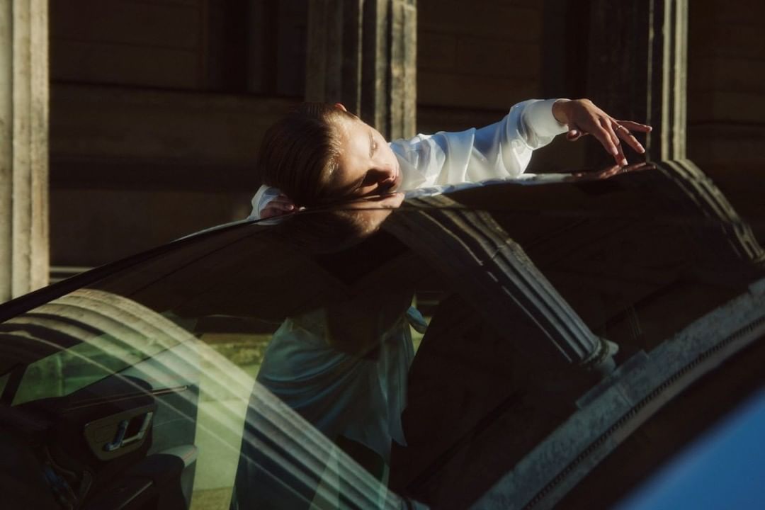 Woman in white shirt leaning on a car roof outdoors with classical columns in background