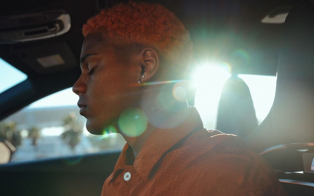 A young adult with orange hair sits inside a car with bright sunlight streaming in, highlighting their profile and creating lens flare.