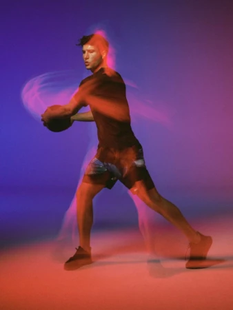 Athletic man in black sportswear dribbles a basketball in a dynamic long exposure studio shot with red and purple gel lighting.