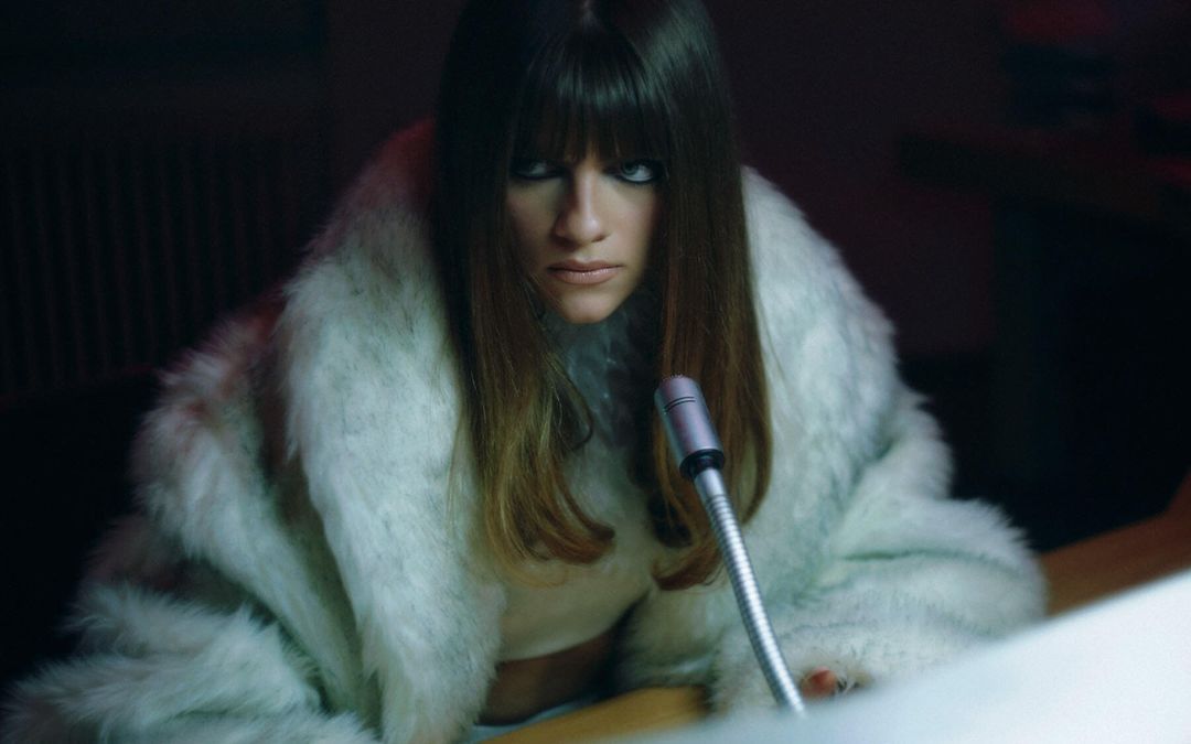 Woman in a white faux fur coat leaning over a desk with a vintage microphone under dramatic low light.