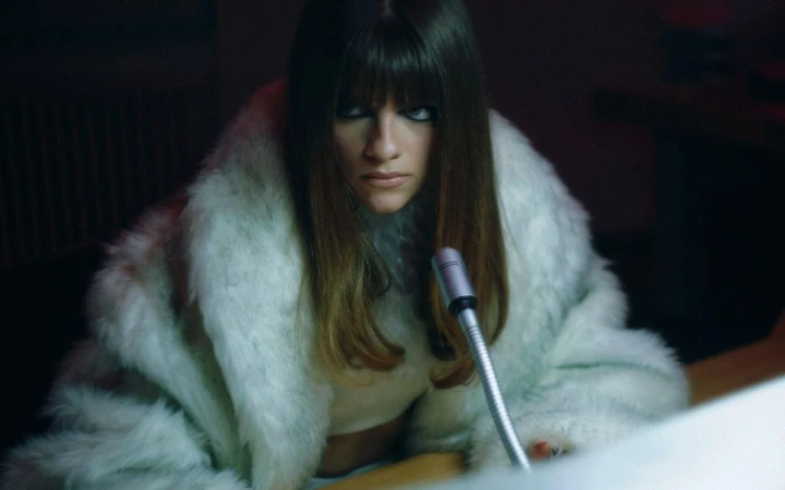 Woman in a white faux fur coat leaning over a desk with a vintage microphone under dramatic low light.