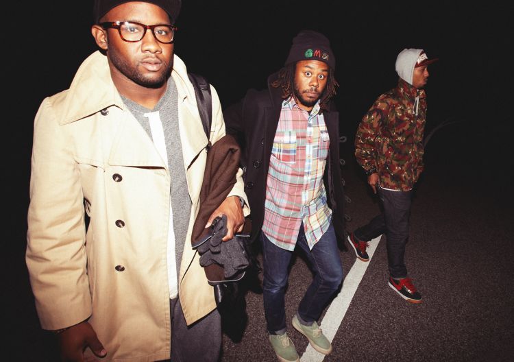 Three Black adult men walking at night. One wears a beige trench coat and glasses. Another wears a plaid shirt. Wide shot.