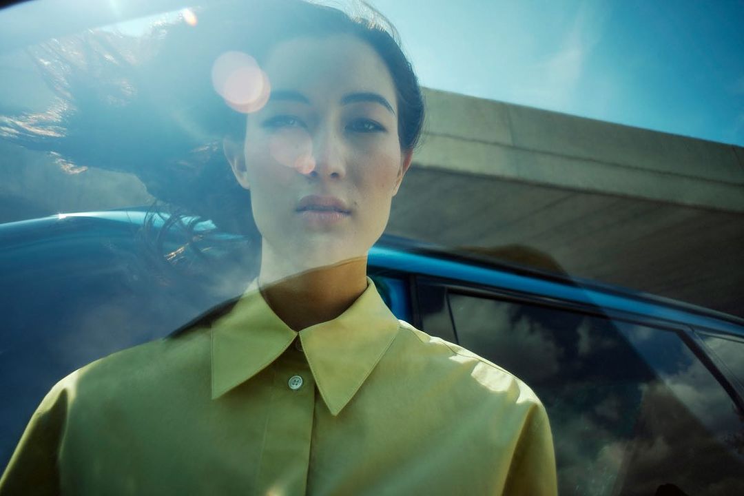Portrait of a woman in a button-down shirt standing in front of a blue car with sunlight flare, under a bright blue sky.