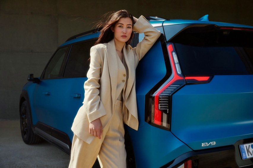 Adult female in a beige suit poses against a blue EV9 car, showcasing automotive fashion and modern vehicle design in a neutral environment.