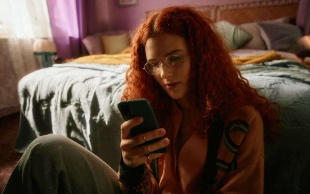Young woman with curly red hair wearing glasses sits indoors using a smartphone. The background includes a neatly made bed.