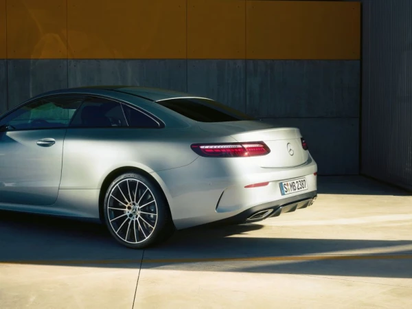 Rear quarter view of a silver Mercedes coupe parked outdoors against a concrete and yellow panel wall with harsh sunlight.