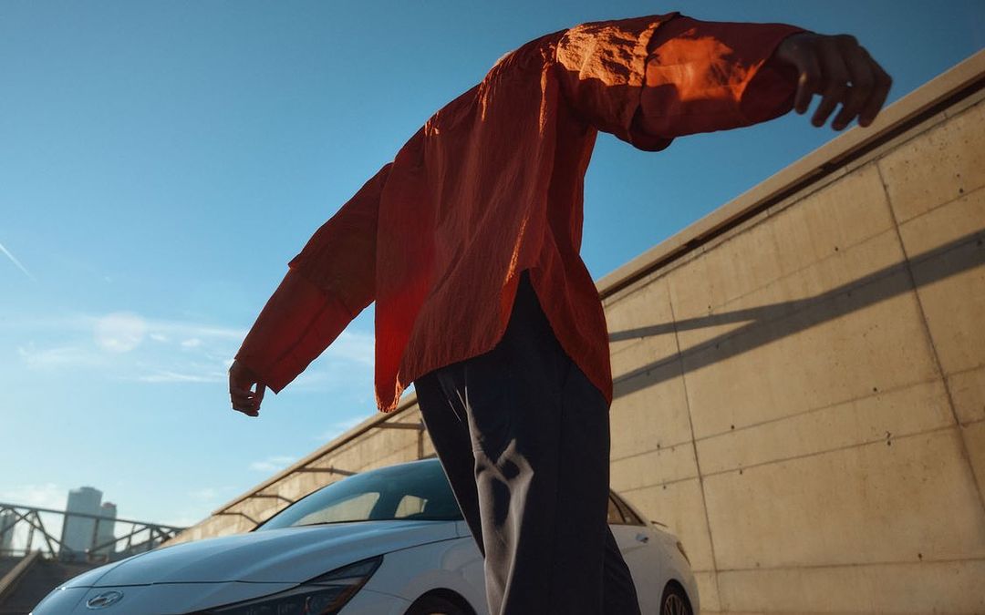 Low angle shot of a person in a red shirt with arms outstretched, standing near a white car, with a concrete wall and blue sky background.