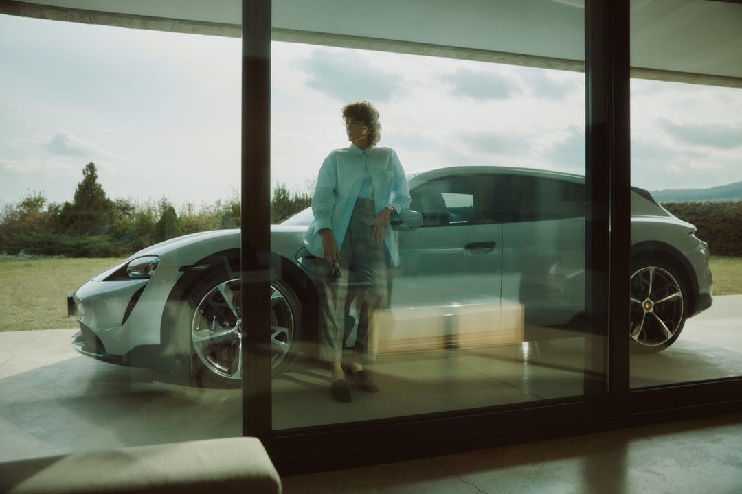 Woman in a light blue shirt stands next to a grey Porsche Taycan Cross Turismo electric vehicle in front of large glass doors.