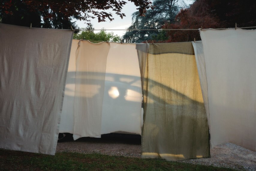 Outdoor photo of white and off-white fabric sheets hanging on a clothesline in a backyard, with trees and the soft glow of sunset in background.