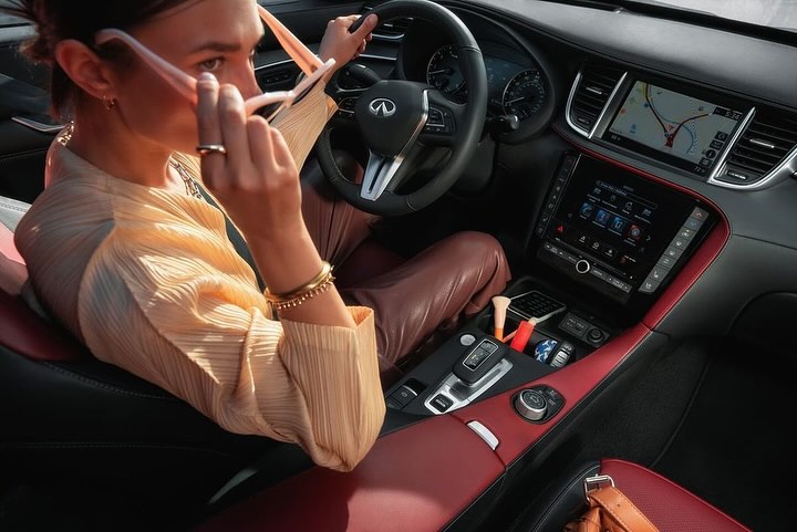 Woman in a yellow blouse holding pink sunglasses in a modern car interior with a red and black dashboard and leather seats.