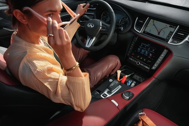 Woman in a yellow blouse holding pink sunglasses in a modern car interior with a red and black dashboard and leather seats.